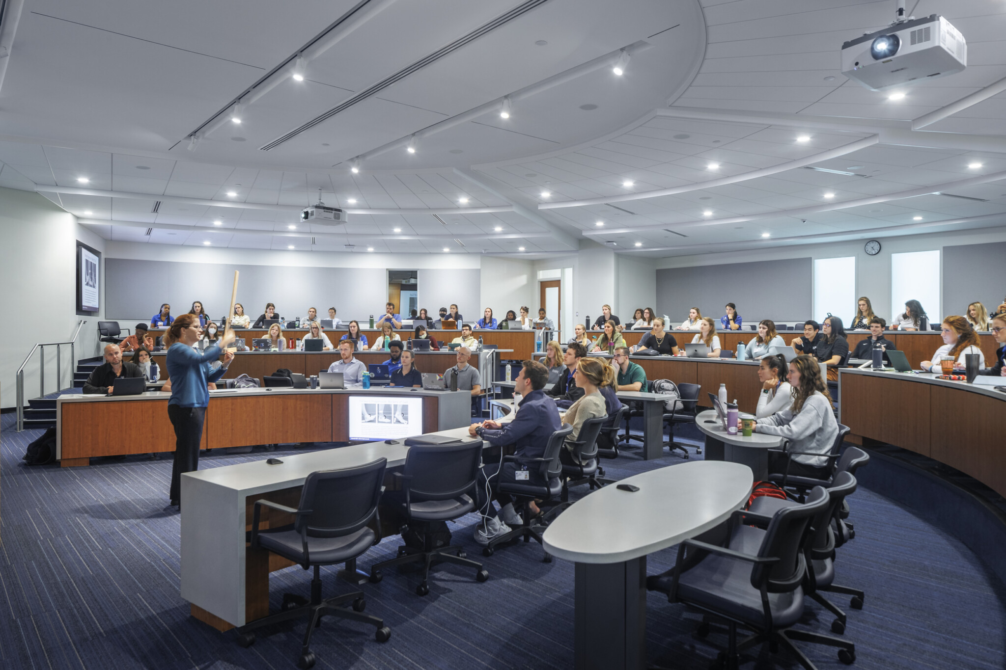 Tiered classroom with multiple rows of seating, integrated AV systems, and a central teaching area.